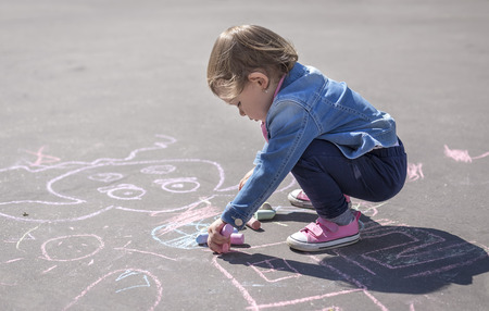 Little beautiful girl in a jeans jacket draws with colored chalks on the playgroundの写真素材