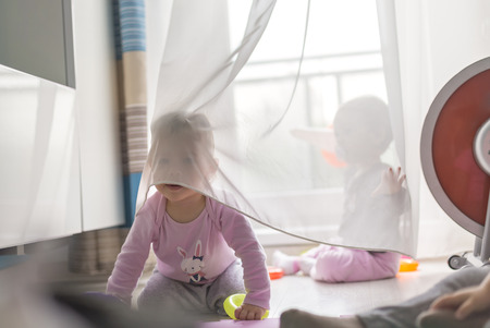 twins play with a curtain at home on the floor, in front of the balcony windowの写真素材