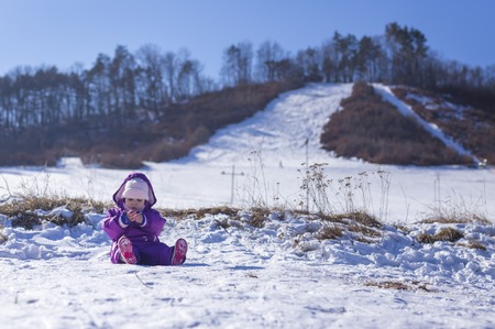Adorable baby in a white snow in the warm suit siting in the snow on a sunny winter day under the hillの写真素材