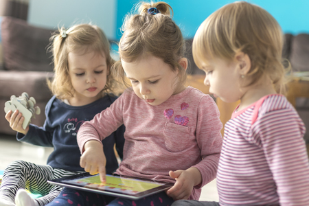 Three little sisters use tablet at home and sit in the middle of toys. Watching something funny.の写真素材