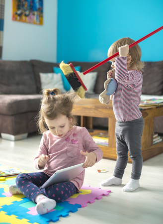 Cute little girl is using a digital tablet  sitting on the floor at home in the middle of toys. Her sister plays with a broom and disturbs her.の写真素材