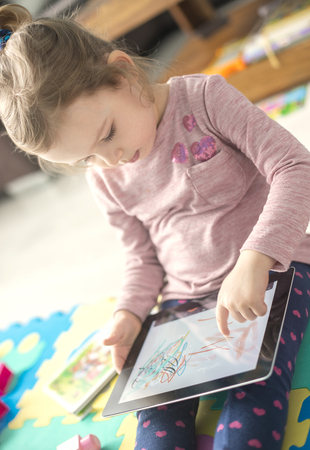 Cute little girl is using a digital tablet  sitting on the floor at home  in the middle of toys.の写真素材