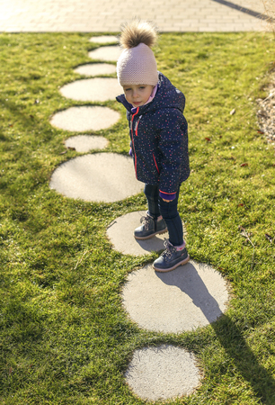 a nice little girl with blue eyes in a winter suit and stocking cap walks along the sidewalk. Concrete rings in grassの写真素材