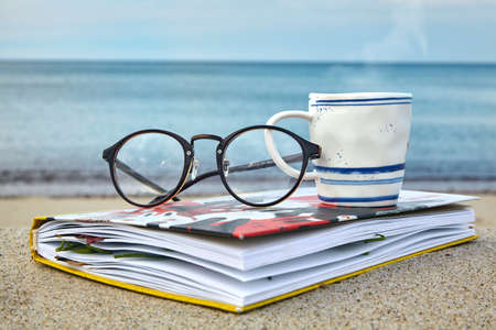 A cup of hot tea, glasses and a notepad with dried leaves on the seashore