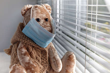 A sad teddy bear in a medical mask looks out the windowの写真素材