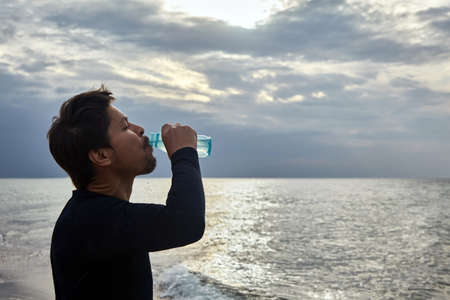 A man drinks water after jogging along the seashore against the backdrop of a sunset skyの写真素材