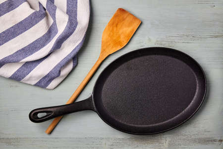 An oval cast iron frying pan with a wooden spatula and a tea towel on a blue wooden table. Template for laying out a dishの写真素材