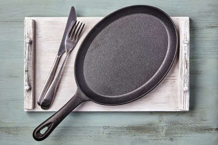 An empty oval cast iron pan on a wooden tray on a light blue kitchen table. Restaurant menu mockupの写真素材