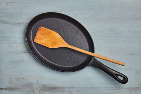 Oval cast iron frying pan with a wooden spatula on a blue wooden table. Menu templateの写真素材