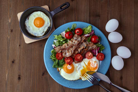 Fried eggs with bacon, lettuce and cherry tomatoes on a blue plateの写真素材