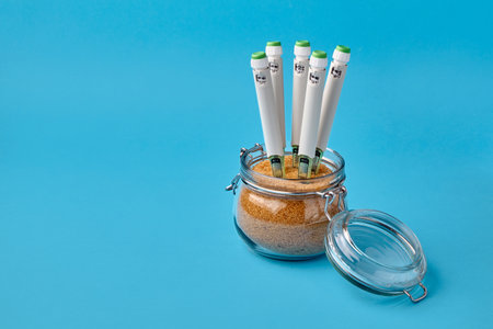 Glass jar with brown cane sugar and insulin syringe pens stuck into it on a blue backgroundの写真素材