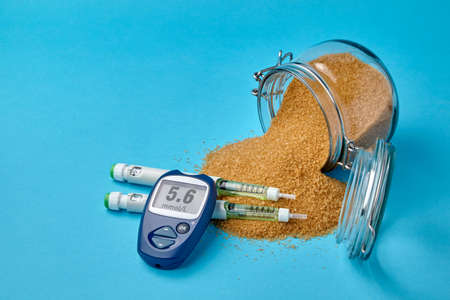 Glass jar with brown cane sugar, insulin syringes and a glucometer on a blue backgroundの写真素材