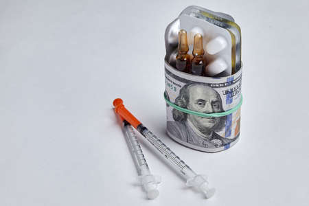 Tablets and ampoules are wrapped in hundred dollar bills, fastened with a rubber band and insulin syringes isolated on a white concrete background. Pandemic and rising drug pricesの写真素材