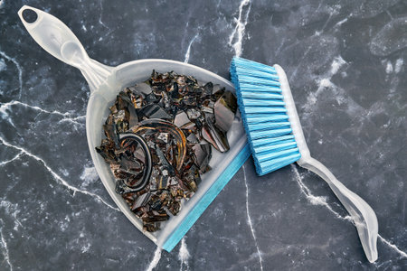 Shards of broken dark glassware on a scoop and a brush on a marble floorの写真素材