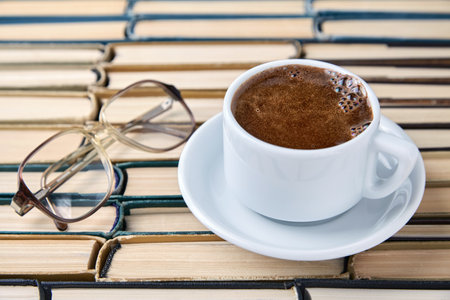 A white cup of fragrant coffee cappuccino and reading glasses against the background of worn old books stacked upside downの写真素材