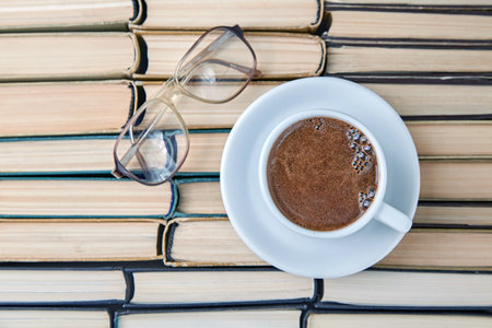 A white cup of fragrant coffee cappuccino and reading glasses against the background of worn old books stacked upside downの写真素材
