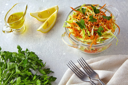 Light summer salad of white cabbage and carrots with sesame and black cumin seeds in a glass bowl on a stone gray table surrounded by ingredients and utensilsの写真素材
