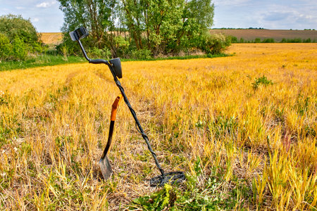 Metal detector and shovel for searching for coins and precious metals against background of yellow field and green forest. Treasure hunting equipmentの写真素材