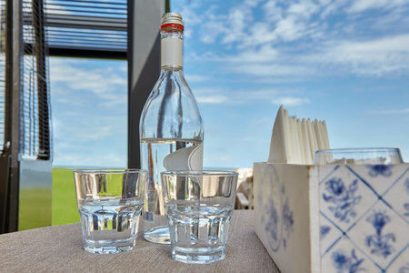 A glass bottle of drinking water and two glasses on the table of an outdoor summer cafe against a background of sky and lawn.の写真素材
