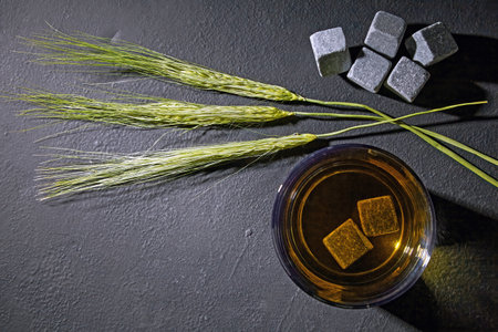 Glass of whiskey with rocks and ears of rye on a black concrete table. Top view with copy space.