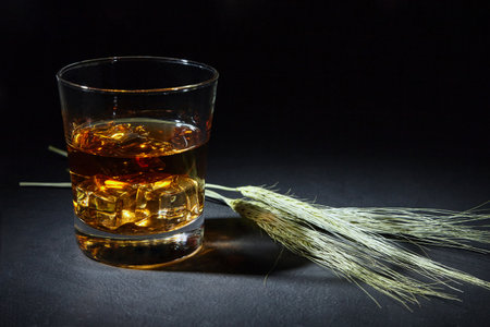 Glass of malt whiskey with ice and ears of wheat on a black concrete table