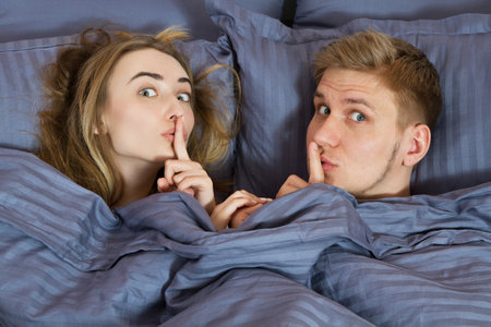 young positive loving couple under blanket in bed and show with fingers the sign of silenceの写真素材