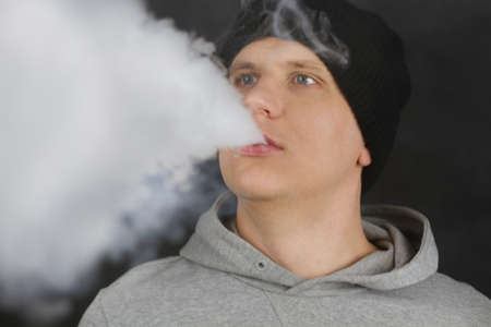 The man smoke an electronic cigarette on the dark background, Men in hoody vaping and releases a...