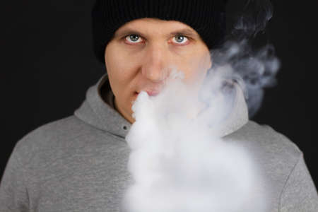 The man smoke an electronic cigarette on the dark background, Men in hoody vaping and releases a...