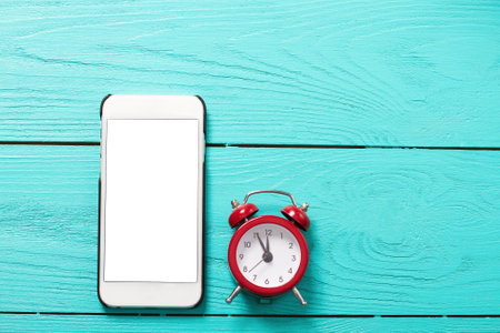 Mobile phone and red retro alarm clock with five minutes to twelve o'clock on blue wooden background. Top view and blank screenの写真素材