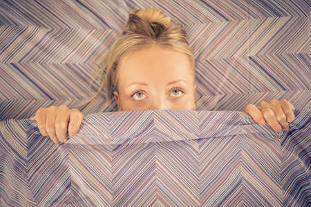 Attractive woman looking from under the blanket - Sleepy girl in bed doesn't want to wake up