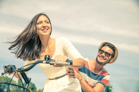 Happy couple riding bikes in the city - Young pretty woman driving bicycle and playful man sitting behind - Portrait of two lovers outdoorsの写真素材