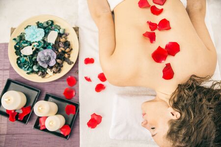 Spa treatment - Beautiful young woman lying on a massage bed with roses and candles and relaxing after a working day - Woman having massage in elagant and relaxing atmosphereの写真素材