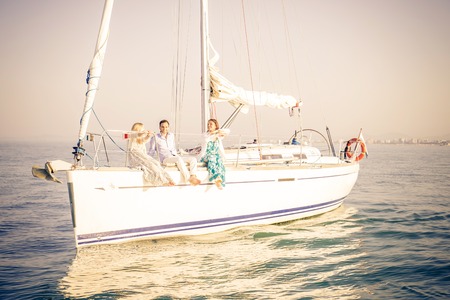 Group of young people sitting on a sailing boat - Two beautiful women and attractive man having fun on a boat while on vacation - Rich people enjoying partyの写真素材