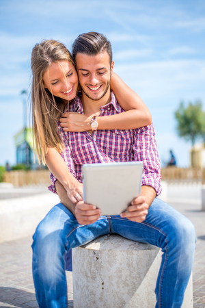 Beautiful couple sitting on a bench outdoors and looking at tablet - Lovers having fun with new technology and shopping onlineの写真素材