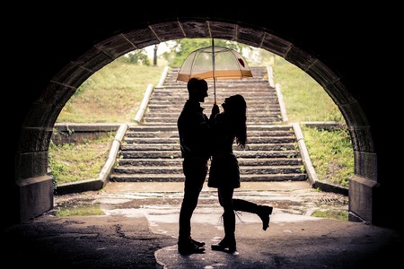 Couple of lovers hugging under a bridge on a rainy day - Silhouettes of man and woman on a romantic date under the rain, laughing and having funの写真素材