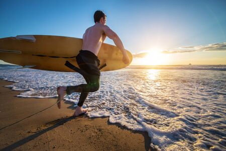 Surfer with board running into the the ocean at sunset - Sportive man going to surf on a tropical beachの写真素材