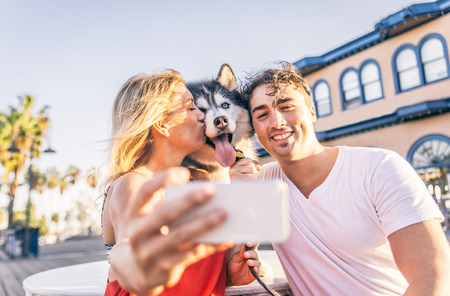 Happy couple taking a selfie with their husky - Woman holding cellphone and taking a funny picture with his familyの写真素材