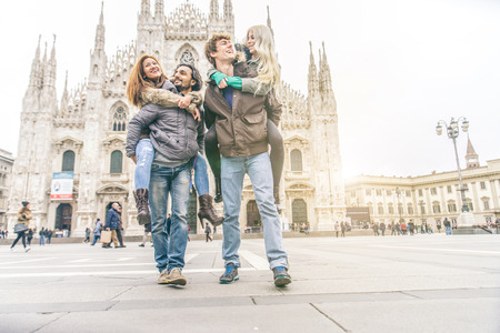 Young friends having fun outdoors - Five students outdoors, men carrying two girls on piggyback - Cheerful young people sightseeing a city, Duomo cathedral in the backgroundの写真素材