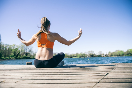 Woman practicing yoga in front of a lake - Girl relaxing and meditating after a workout sessionの写真素材