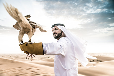 Arabic man with traditional emirates clothes walking in the desert with his falcon bird