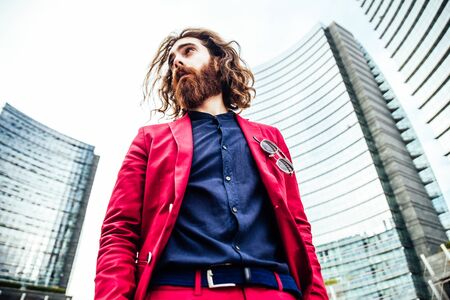 Fashion portrait of a young hipster man walking outdoors with a red formal suit - Unusual businessman going to workの写真素材