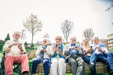 Group of senior people learning to use modern technologies - Mature seniors sitting on bench in a park and staring at cellphonesの写真素材