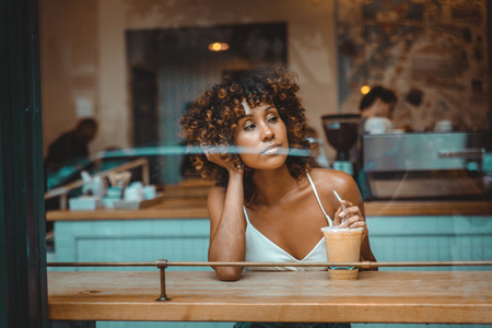 Beautiful afro woman sitting in a coffehouse - Pretty middle aged girl drinking coffee in a bar, reflection effect from window glassの写真素材