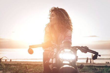 Beautiful girl having fun driving her custom cafe racer motorcycle, enjoying the sunset on the beachの写真素材