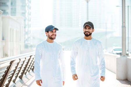 Two young men going out in Dubai. Friends wearing the kandura traditional male outfit and baseball hat in Marinaの写真素材
