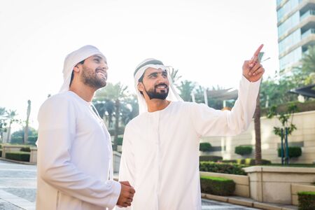 Middle-eastern young adults wearing kandora walking outdoors in Dubai - Two businessmen meeting outdoorsの写真素材