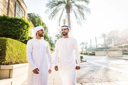 Middle-eastern young adults wearing kandora walking outdoors in Dubai - Two businessmen meeting...