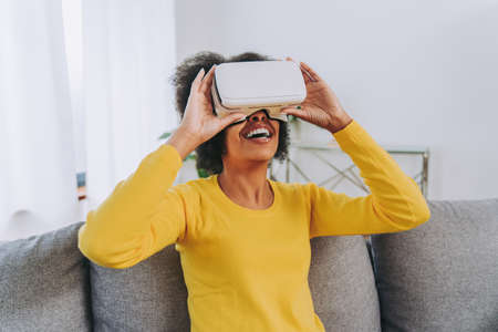Beautiful woman playing at home with VR headset - Black woman lying down on sofa while interacting...