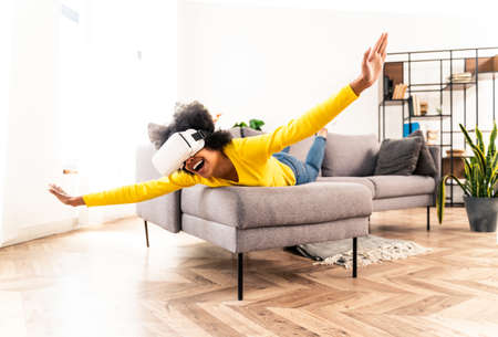 Beautiful woman playing at home with VR headset - Black woman lying down on sofa while interacting with augmented reality technologyの写真素材