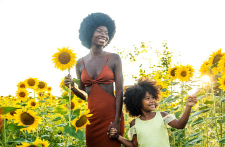 Beautiful afro-american mom and daughter palying and having fun in a sunflowers fieldの写真素材
